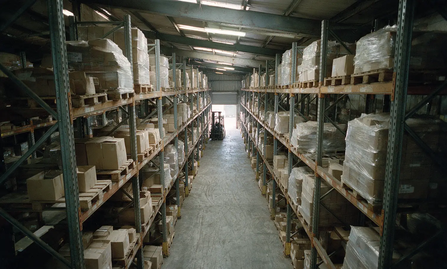 A narrow aisle between tall pallet racking in an industrial valve warehouse, cartons and wrapped pallets on every level
